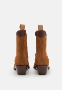 Stivaletti in suede marrone con un accento in pelle liscia, pannelli elastici sui lati e tacco in legno basso e impilato. Caratteristici nastri di fissaggio sul retro.