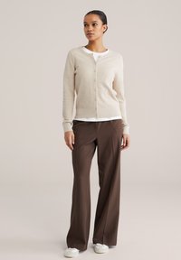 Béžový pletený sveter s textúrovaným vzorom, zapínaný na gombíky vpredu, s rebrovaným lemom, doplnený o hnedé nohavice s širokými nohavicami a biele tenisky.