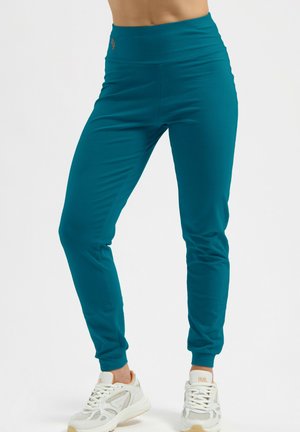 Frau trägt türkisblaue Leggings mit hohem Bund und weiße Sneakers, steht vor weißem Hintergrund, Beine leicht auseinander.