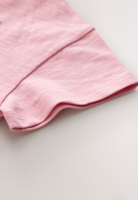 Detail růžové košile s krátkým rukávem, zobrazující strukturu látky a šitý lem na světlém pozadí.