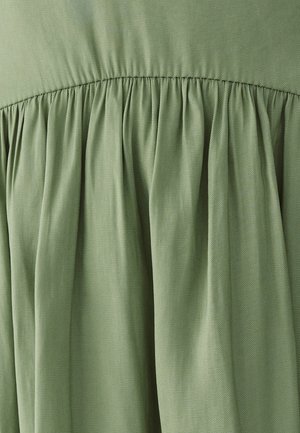 Lichtgroene stof met diagonale weefstructuur, verzameld in zachte plooien onder een gestikte naad.