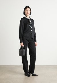 Blouse noire à boutons avec un motif texturé, associée à un pantalon noir ajusté et un petit sac à main noir ; accentuée avec une écharpe noir et blanche.