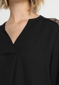 Blusa nera con scollatura a V, dettagli in pizzo sulle spalle e una texture di tessuto liscia. Un design che enfatizza semplicità ed eleganza.