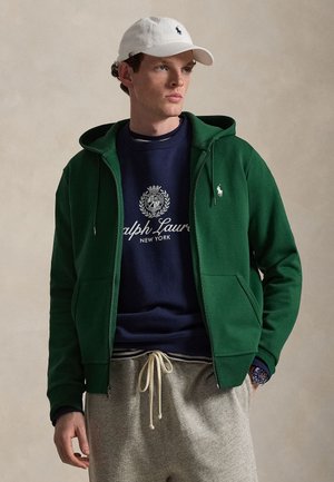 Mladý muž s bielou čiapkou, zelenou mikinou na zips, modrou mikinou Ralph Lauren a sivými teplákmi, stojaci s jednou rukou v vrecku.