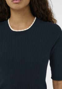 Vrouw in een marineblauw ribgebreid shirt met korte mouwen en een witte scalloped halslijn, weergegeven van schouders tot halverwege de romp.