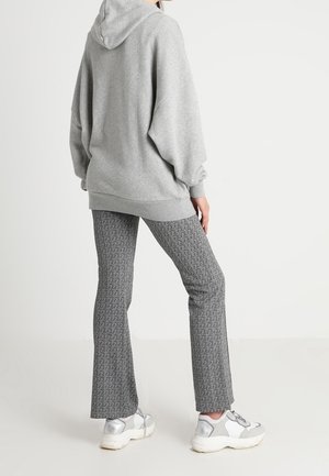 Personne debout, portant un sweat à capuche gris ample, un pantalon évasé gris à motifs et des baskets épaisses blanches et grises sur un fond uni.