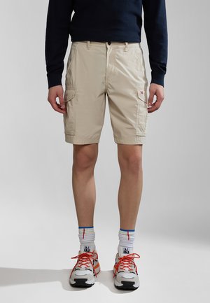 Pantaloni cargo beige con tasche laterali, vita piatta e chiusura con bottone. Abbinati a calzini bianchi a fantasia e sneakers multicolori.