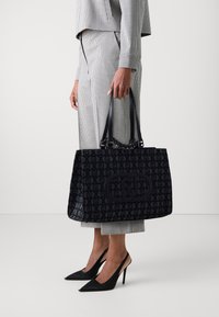 Borsa tote nera con motivo, dotata di due manici e tessuto testurizzato; presenta un grande patch con il logo sul davanti. Indossata con un abbigliamento a scacchi.