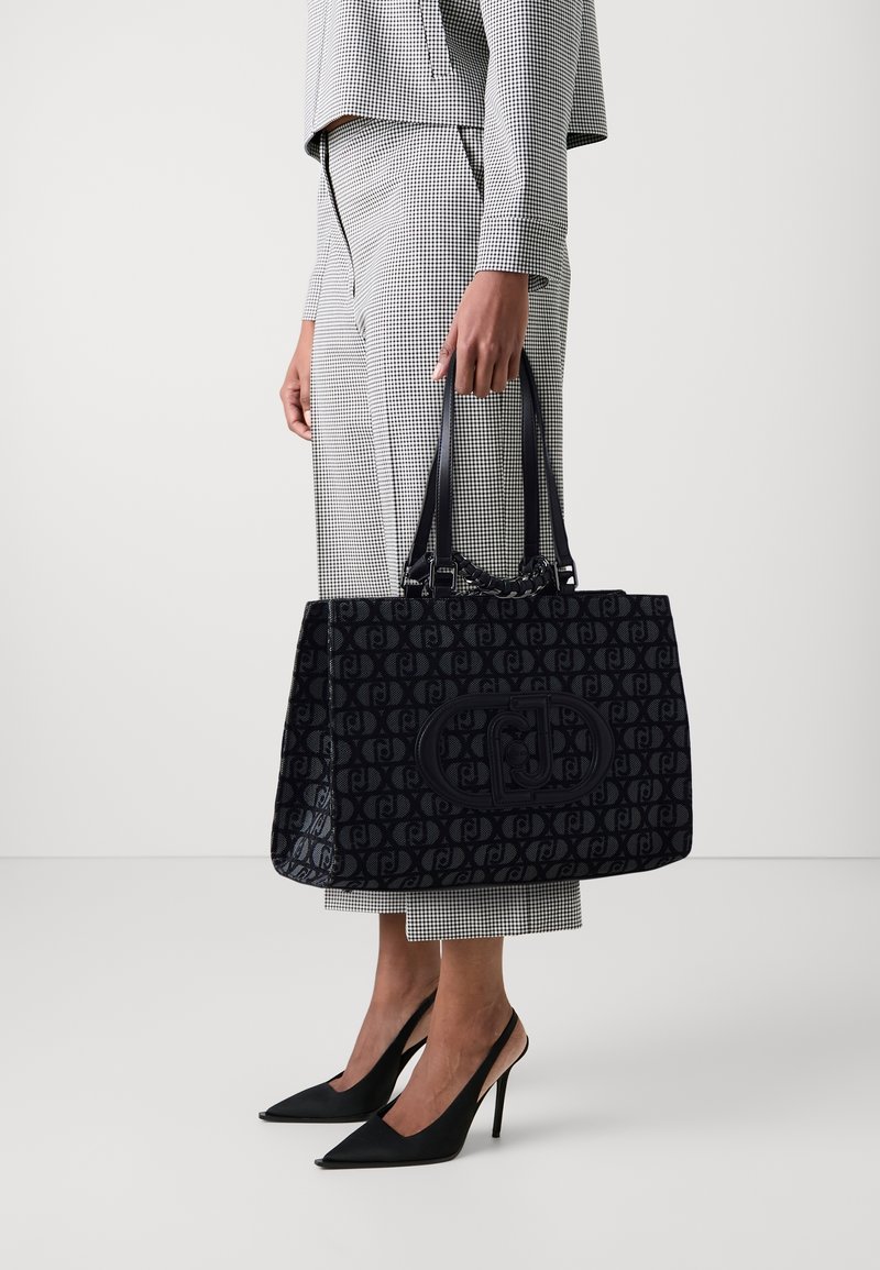 Borsa tote nera con motivo, dotata di due manici e tessuto testurizzato; presenta un grande patch con il logo sul davanti. Indossata con un abbigliamento a scacchi.