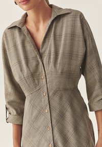 Abito camicia a quadri marrone con chiusura frontale a bottoni, colletto e maniche a tre quarti. Presenta un tessuto strutturato con motivi a quadri variegati.