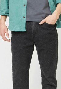Jeans in denim nero con vestibilità dritta, trama sottile e tasche frontali. Abbinati a una camicia teal a bottoni e una maglietta grigia sotto.