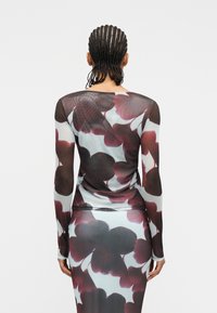 Άτομο με πλεγμένα μαλλιά, φορώντας διάφανο τοπ με μακριά μανίκια και φούστα με μεγάλα σκοτεινά κόκκινα και μαύρα λουλουδένια σχέδια, γυρισμένο με την πλάτη σε λευκό φόντο.