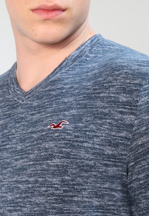Zbliżenie osoby noszącej niebieską teksturowaną koszulkę z dekoltem w serek i małym czerwonym logo ptaka na piersi.