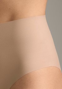 Prim-plan al taliei și șoldului unei persoane care poartă lenjerie nud fără cusături, cu talie înaltă, pe un fundal simplu.