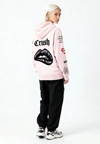 Felpa con cappuccio rosa con tasca frontale, caratterizzata da una grafica di labbra nere e testo sul retro. Abbinata a pantaloni cargo neri e sneaker.