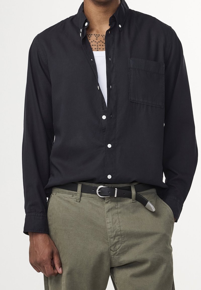 Homme portant une chemise noire boutonnée sur un sous-vêtement blanc, pantalon vert olive et ceinture noire avec boucle argentée, main dans la poche.