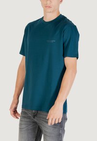 T-shirt in cotone turchese con collo rotondo, maniche corte e logo discreto sul petto sinistro; abbinato a jeans in denim grigi.