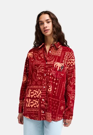 Vrouw met lang golvend haar, gekleed in een rood en oranje patroon overhemd met knopen en lichtblauwe spijkerbroek, staand tegen een effen achtergrond.