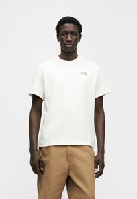 Λευκό βαμβακερό t-shirt με κοντά μανίκια και λαιμόκοψη σε στρογγυλή λαιμόκοψη, με μικρό καφέ λογότυπο στο αριστερό στήθος, σε συνδυασμό με μπεζ παντελόνια.