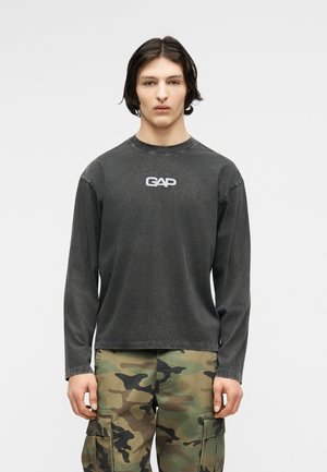 Νεαρό άτομο με σκούρα μαλλιά μέχρι τους ώμους, που φοράει μαύρο μακρυμάνικο μπλουζάκι GAP και παραλλαγμένες παντελόνες cargo, στέκεται μπροστά σε ένα μονόχρωμο φόντο.