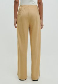 Lichte beige wijde broek van stof met een gladde textuur. Heeft een hoge taille en subtiele zijnaad, en gaat tot aan de enkels.