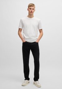 Witte katoenen t-shirt met een ronde hals, gecombineerd met zwarte broek. Het model draagt beige sneakers met roomkleurige en bruine accenten. Eenvoudig ontwerp.