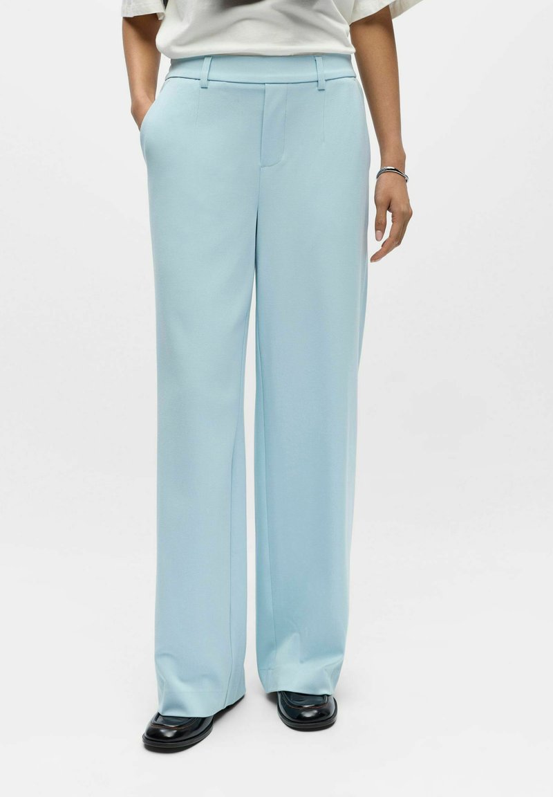 Pantalon bleu clair à jambes larges avec une texture lisse, doté de poches avant et d'une coupe frontale plate. Assorti à des chaussures noires.