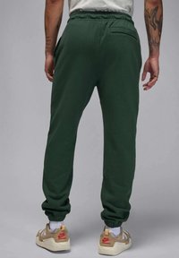 Pantaloni verdi in felpa con vita e polsini elasticizzati. Presentano una tasca posteriore. Indossati con sneaker beige e rosse. Texture liscia, simile al cotone.