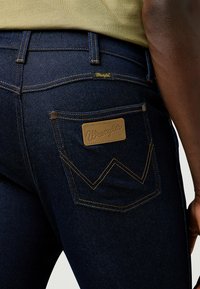 Jean en denim bleu foncé avec une étiquette en cuir sur la poche arrière portant le logo "Wrangler" en relief. Coutures jaunes visibles et design classique des poches.