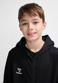 Schwarze Zip-Hoodie aus weichem Stoff, mit einer großen Kapuze und einem weißen Logo auf der Brust. Schlichte Design ohne Muster oder Verzierungen.