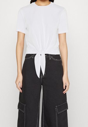 Žena v bílém krátkém topu s uzlem vpředu, doplněném o černé široké kalhoty s vysokým pasem, velkými bočními kapsami a bílým prošíváním.