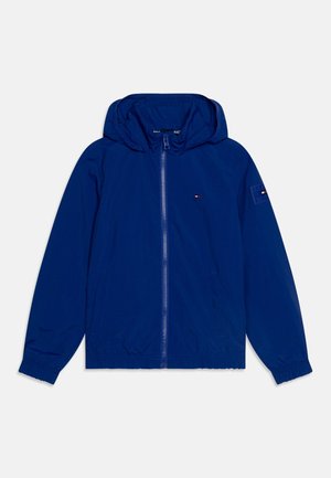 Chaqueta ligera azul con capucha, cremallera frontal, puños y dobladillo elásticos, y parche de logo en la manga. Textura suave, diseño casual.