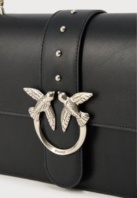 Μαύρη δερμάτινη τσάντα χειρός με ασημένιο κυκλικό διακοσμητικό στοιχείο με δύο μοτίβα πουλιών, διακοσμητική λεπτομέρεια με καρφιά κατά μήκος του λουριού και λεία υφή.