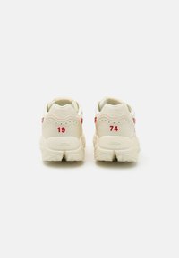 Sneakers med en kremfarget overdel, røde detaljer, og preget tallene 19 og 74 på hælen. Teksturert såle og logo vist på baksiden.