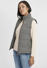 Graue gesteppte Weste mit hohem Kragen, Frontreißverschluss und Druckknöpfen. Trägt man über einem hellbeigen Pullover. Verfügt über ein kleines Logopatch.