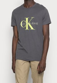 Šedé bavlněné tričko s velkým žlutým logem "CK" a nápisem "CALVIN KLEIN JEANS", krátké rukávy a kulatý krk.
