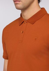 Koszulka polo z krótkim rękawem w kolorze rdzawego pomarańczu, wykonana z miękkiego materiału, z ściągaczem przy kołnierzyku, zapięciem na trzy guziki i subtelnym haftem logo.