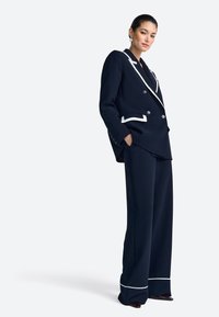 Blazer azul marino cruzado con ribete blanco, combinado con pantalones de pierna ancha, que presenta un diseño elegante y detalles sutiles de textura.