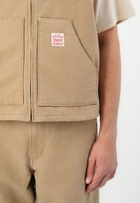 Beige werkkleding vest met twee voorzakken, een rood Levi Strauss & Co. label op de rechterzak, gedragen over een beige t-shirt.