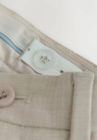 Svetlo bež tkanina z teksturirano površino, z modrim elastičnim trakom, belim gumbom za zapiranje in ravnim bež pasom z vidnimi šivi.