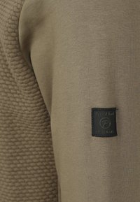 Olijfgroen sweatshirt met een textuurpaneel aan één kant, voorzien van een zwart merklabel op de onderste zoom. Gladde stof, casual ontwerp.