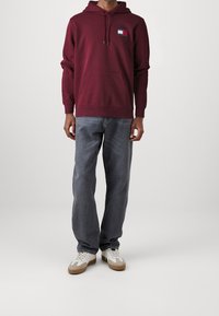 Felpa con cappuccio bordeaux con tasca frontale e logo, abbinata a jeans grigi chiari ampi e sneakers bianche con dettagli marroni.