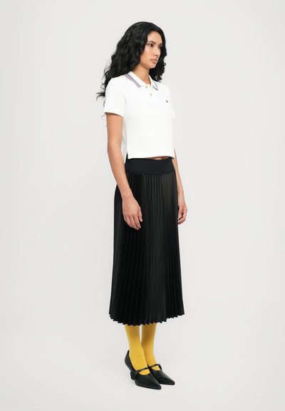 Hvid cropped polo shirt med en flerfarvet krave, parret med en sort plisseret nederdel, gule tights og sorte spidse sko.