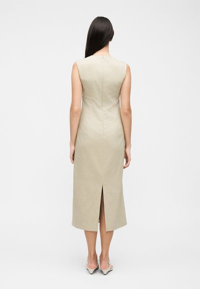 Γυναίκα με μακριά σκούρα μαλλιά που φοράει μπεζ μίντι φόρεμα χωρίς μανίκια με σχίσιμο στο κέντρο της πλάτης και μπεζ τακούνια, στέκεται μπροστά σε ένα λευκό απλό φόντο.