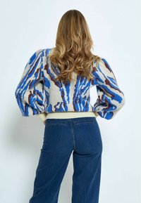 Strickpullover mit einem blauen, braunen und beigen abstrakten Muster, puffigen Ärmeln und einem gerippten Saum, kombiniert mit dunkelblauen weiten Jeans.