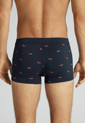 Homme portant un boxer bleu marine orné de petits motifs de teckels rouges, debout de dos face à la caméra sur un fond uni.