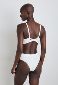 Hvit bralette og høytlivde truser laget av myk stoff, med justerbare stropper og en subtil tekstur, samt lukking på baksiden.