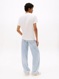 Witte katoenen t-shirt met een ronde hals, gedragen met lichtblauwe wijde spijkerjeans. Model staat met de rug naar de camera.