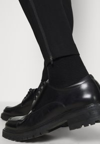 Zapatos de cuero negro con superficie lisa, punta redondeada y suela de goma gruesa; combinados con calcetines negros acanalados y pantalones negros ajustados con detalle de cremallera.