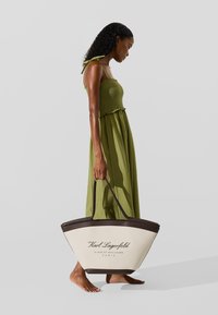 Suuri kermainen olkalaukku, jossa tummanruskeat yksityiskohdat ja brändin nimi painettuna mustalla värillä. Mallilla on päällään vihreä mekko, jossa on rypytetty yläosa.